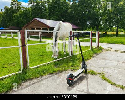 Pferd in der Arena. Im Stall ist ein schönes weißes Pferd. Ein Elektroroller steht in der Nähe der Pferdearena. Tier- und Elektrotransport. Vergangenheit Stockfoto