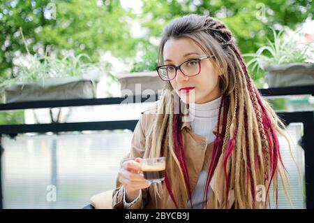 Positive smart Student Mädchen mit langen Dreadlocks ruht in einer Straße Coffee-Shop mit transparenten Tasse Espresso. Stockfoto