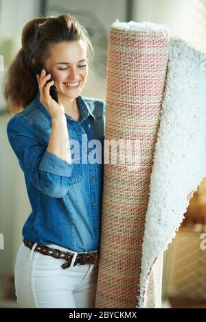 Lächelnde moderne Frau in Jeans-Hemd und weiße Hose mit weißem Teppich mit einem Smartphone im modernen Haus an sonnigen Tag. Stockfoto