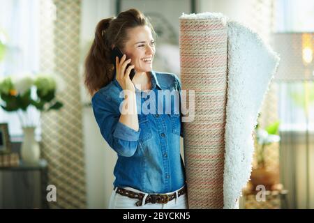 Happy Modern Mittelalterhausfrau im Jeans-Hemd und weiße Hose mit weißem Teppich mit einem Smartphone im modernen Wohnzimmer an sonnigen Tag. Stockfoto