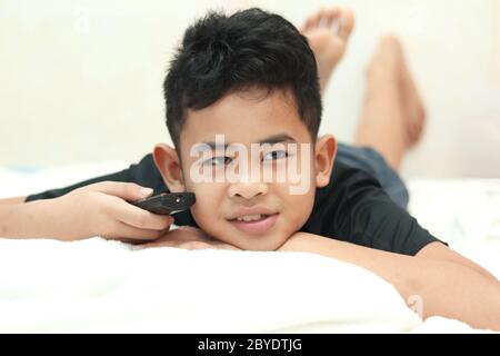 Niedliches kleines asiatisches Kind, das im Bett liegt und die fernbedienung hält und im Schlafzimmer zu Hause Fernsehen schaut, Unterhaltung für Kinder Stockfoto