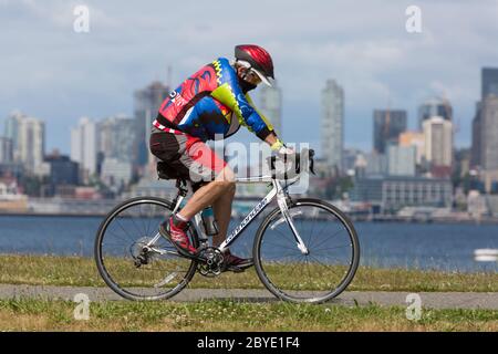 Ein Radfahrer mit einer schützenden Gesichtsmaske fährt an einem sonnigen Nachmittag in Seattle, Washington, entlang des Alki Trail. Stockfoto
