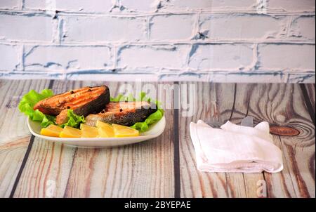 Zwei gegrillte rosafarbene Lachssteaks liegen auf einer weißen Keramikplatte, die mit Zitronenscheiben und einem Salatblatt neben einer weißen Serviette mit Besteck verziert ist, dem WH Stockfoto