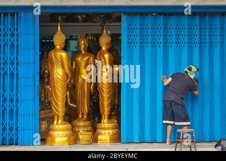 Ein Mitarbeiter eines Ladens in Bamrung Muang Rd., Bangkok, Thailand, Reinigung der Türfenläden, Buddha-Statuen zum Verkauf hinter der halb geöffneten Klappe gesehen Stockfoto