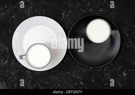 Minimalistische flache Lay von zwei Tassen Milch stehen auf schwarzen und weißen Tellern, ähnlich dem Yin Yang Konzept auf einem strukturierten schwarzen Hintergrund. Stockfoto