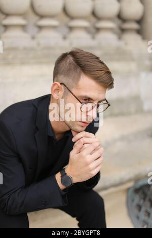 Junger europäischer Mann in einer Brille auf dem Bürgersteig sitzend, der sich auf einem Betongeländer stützt. Stockfoto
