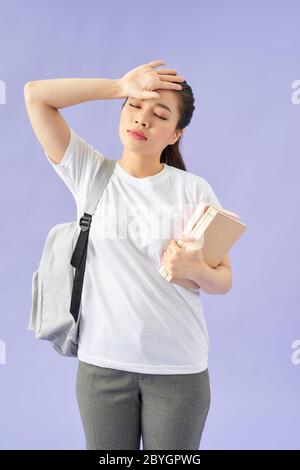 Müde junge Frau Studentin mit Rucksack auf lila Hintergrund. Halten Sie Bücher mit geschlossenen Augen Hand auf den Kopf Stockfoto