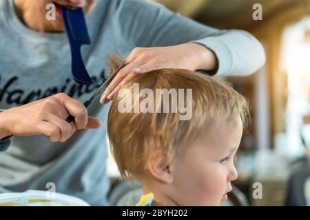 Close-up junge Erwachsene kaukasische Mutter machen Haarschnitt fot niedlich liebenswert Sohn Kleinkind Junge zu Hause durch Quarantäne und Sperrung. Mutter schneiden Haare des Kindes Stockfoto