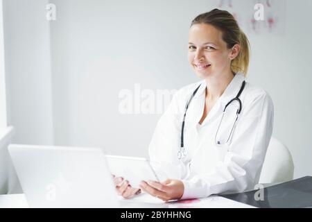 Die junge, blonde Ärztin sitzt in ihrer Praxis, arbeitet auf ihrem Tablet und schaut sich die Untersuchungsergebnisse an Stockfoto