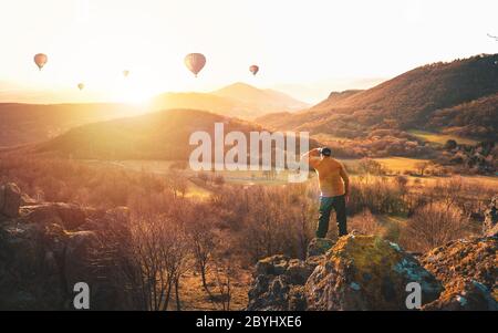Abenteuerlicher Mann steht auf einem Berg und genießt eine schöne Aussicht während viele Ballons. Das aktive Lifestyle-Wochenende. Luftballons fliegen Stockfoto