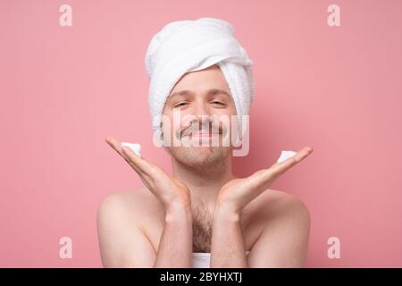 Schöner lustiger Mann mit Handtuch auf dem Kopf, der seine Haut mit Schaum tut Spa-Hautpflege. Studio auf rosa Wand gedreht. Stockfoto