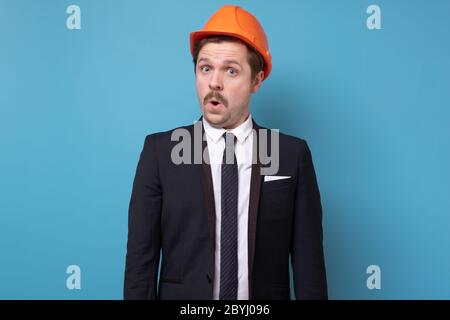Junge Bauarbeiter in specia-Hut und Anzug ist schockiert über Nachrichten auf blauem Hintergrund Stockfoto