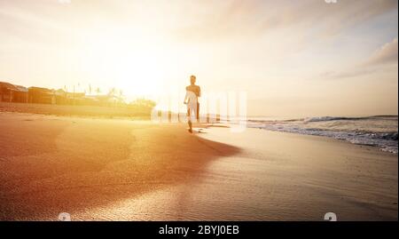 Bild von feinem Mann beim Training am Morgen am Strand entlang laufen. Jogging an der Küste entlang mit schönem orangefarbenen Sonnenaufgang über dem Meer im Hintergrund Stockfoto