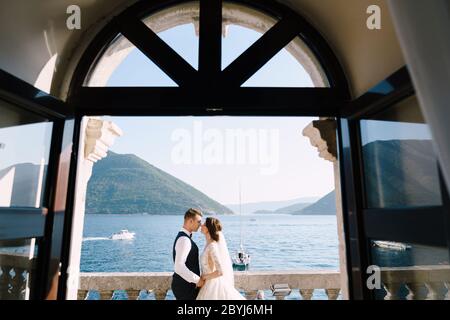 Die Braut und der Bräutigam stehen auf dem Balkon des Hotels mit Blick aufs Meer, ein Blick durch das offene alte Fenster. Fine-Art Hochzeitsfoto in Stockfoto