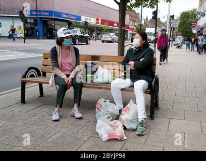 Zwei koreanische Frauen, die weniger als 2 Meter voneinander entfernt sind, tragen Gesichtsmasken und sitzen auf einer Bank in New Malden High Street und unterhalten sich miteinander. Stockfoto