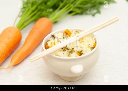Vegetarische / vegane Küche - Rohe Karotten mit Blättern und eine Schüssel gekochten Reis mit Karotten. Essstäbchen oben auf dem Behälter. Stockfoto