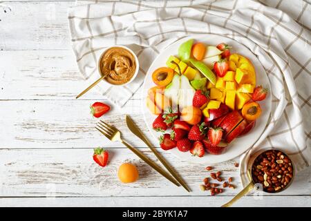 Buntes Obst-Dessert: Erdbeeren, Mango, Äpfel, Birnen, Aprikosen, Erdnussbutter und Erdnüsse auf einem weißen Teller auf einer weißen Holztafel Stockfoto