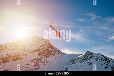 Roter Hubschrauber, der während eines Wintersonnenvergangs über die schneebedeckten Berge der Alpen fliegt. Luftlandschaft Stockfoto