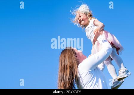 Mama hebt Tochter hoch in blauen Himmel. Glückliche Mutter und Mädchen spielen Spaß im Park im Sommer zusammen auf dem grünen Gras. Familienleben. Stockfoto