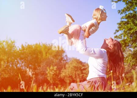 Mama hebt Tochter hoch in blauen Himmel. Glückliche Mutter und Mädchen spielen Spaß im Park im Sommer zusammen auf dem grünen Gras. Familienleben. Stockfoto