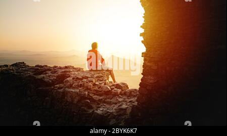 Professioneller Mann sitzt auf Burgruinen in aktiver Kleidung auf dem Gipfel des Berges ruhen und bewundern grüne Hügel und atemberaubende Landschaft Stockfoto