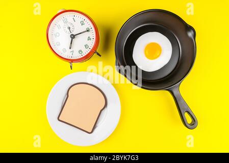 Bratpfanne mit Spiegelei und Wecker isoliert auf gelbem Hintergrund von oben gesehen.Konzept des Frühstücks beim Rennen gegen die Zeit. Stockfoto