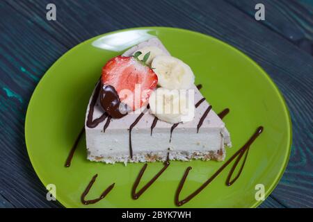 Käsekuchen auf einem Teller. Garniert mit Erdbeeren, Bananen und Schokolade. Auf gebürsteten Kiefernholzplatten schwarz und grün lackiert. Stockfoto