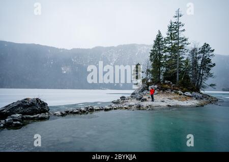 Mann in roter Jacke mit Blick auf gefrorenen See. Kleine Insel mit Bäumen am Eibsee Bayern Zugspitze. Schöner Wintertag Stockfoto