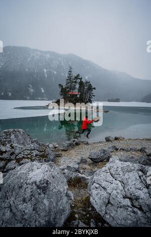 Mann in roter Jacke mit Blick auf gefrorenen See. Kleine Insel mit Bäumen am Eibsee Bayern Zugspitze. Schöner Wintertag Stockfoto