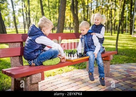 Kleine Schulkinder spielen Schach im Park auf der Bank. Sommerferien, intellektuelle Spiele. Schachclub für Kinder. Glückliche Kindheit und Kinder Stockfoto