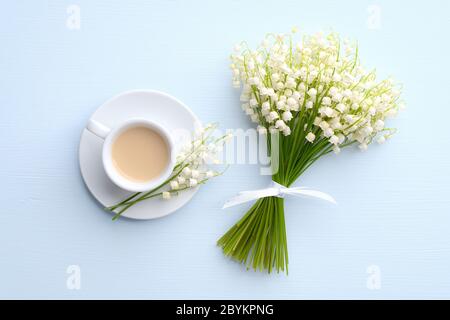 Kaffeebecher mit Blumenstrauß Maiglöckchen auf blauem Hintergrund, Blick von oben. Guten Morgen Kaffee, glücklich Mutter Tag Konzept. Stockfoto