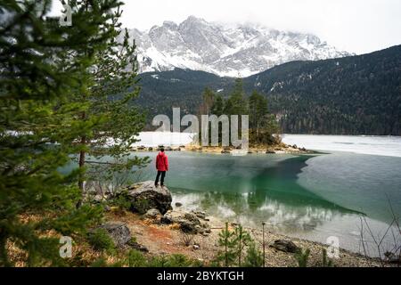 Mann in roter Jacke mit Blick auf gefrorenen See. Kleine Insel mit Bäumen am Eibsee Bayern Zugspitze. Schöner Wintertag Stockfoto