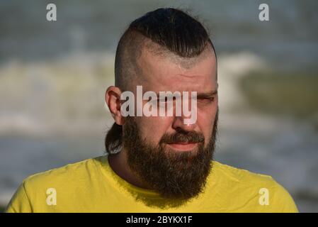 Ein junger Mann, der ein gelbes T-Shirt und Shorts trägt. Mann in einem gelben T-Shirt mit Bart auf dem Hintergrund des Meeres. Stockfoto