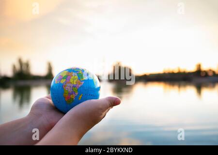 Detail der Hände der Person, die kleinen Ball der Welt vor einem See bei Sonnenuntergang hält. Selektiver Fokus. Konzept der Reise und Pflege des Planeten. Stockfoto