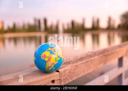 Kleine Weltkugel, die auf dem hölzernen Zaun eines großen Sees aufgestellt wird. Reisekonzept und Pflege des Planeten. Stockfoto