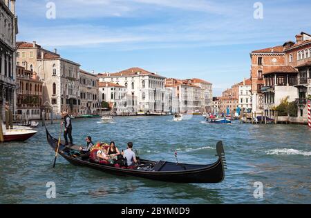 Familie mit einem kleinen Baby bei einer Gondelfahrt auf einem abgehackten Canal Grande in Venedig, Italien, sonnenbeschienene Gebäude im Hintergrund unter blauem Himmel Stockfoto