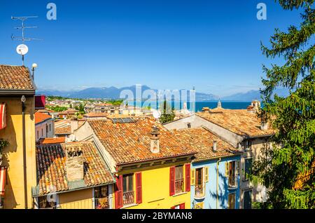 Luftpanorama von Desenzano del Garda Stadt mit rotem Ziegeldach von bunten Gebäuden mit Fensterläden, Gardasee Wasser, Bergkette, blauer Himmel Hintergrund, Lombardei, Norditalien Stockfoto