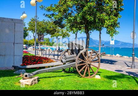 Kanone auf grünem Rasen im historischen Zentrum von Desenzano del Garda Stadt mit Blick auf Gardasee Promenade Böschung und Bergkette, blauer Himmel Hintergrund, Lombardei, Norditalien Stockfoto