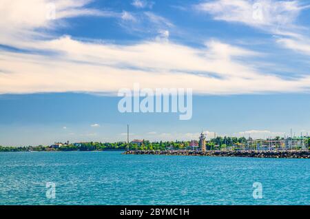 Gardasee azurblaue türkisfarbene Wasseroberfläche mit Blick auf Leuchtturm auf Steinpier Maulwurf und Halbinsel Sirmione, blauer Himmel weiße Wolken Hintergrund, Desenzano del Garda Stadt, Lombardei, Norditalien Stockfoto