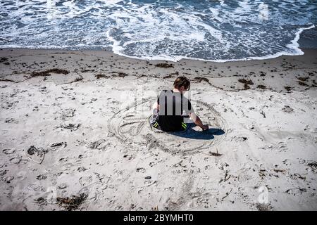 Dieser Mann sitzt am Strand und zieht im Sand seinen ,Kreis seines Lebens'. Die großen Herzen, die kleinen Kreise und die Bedeutung werden weggespült. Stockfoto