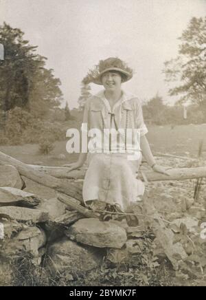 Antike c1920 Fotografie, junge Frau in 1920er Jahren Mode sitzt auf einem Holz und Stein Zaun. Genaue Lage unbekannt, wahrscheinlich New Hampshire, USA. QUELLE: ORIGINALFOTO Stockfoto