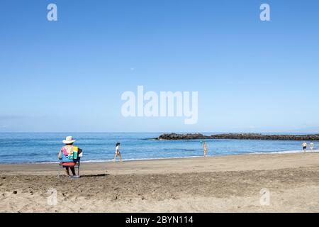 Frau in Strohhut sitzt auf einem tragbaren Stuhl am Strand Playa del Duque, Costa Adeje, Teneriffa, Kanarische Inseln, Spanien Stockfoto