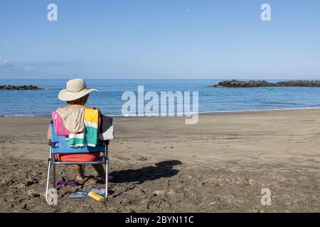 Frau in Strohhut sitzt auf einem tragbaren Stuhl am Strand Playa del Duque, Costa Adeje, Teneriffa, Kanarische Inseln, Spanien Stockfoto