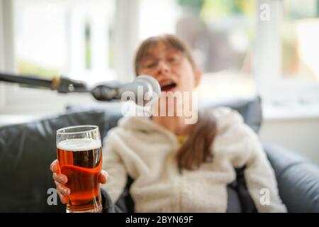 Isolierte junge Frau, die zu Hause Bier trinkt und ins Mikrofon singt. Betrunkene Frau, die ein Bier hält und sich amüsiert! Trinken von überschüssigem Alkohol. Stockfoto