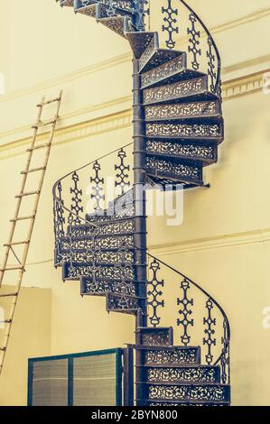 Vintage schwarz geschmiedete Wendeltreppe und Bambusleiter lehnen sich an gelbe Wand Stockfoto