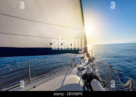 Segeln bei ruhigem Wetter mit Sonne. Blick vom Deck der Yacht auf den Bug und die Segel. Segelboot mit aufgesetzten Segeln, die im offenen Meer gleiten. Griechenland, Europa Stockfoto