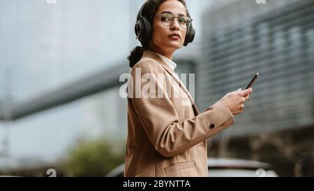Geschäftsfrau mit Kopfhörer, die mit einem Handy in der Hand auf der Straße unterwegs ist. Weibliche Führungskraft über die Straße. Stockfoto
