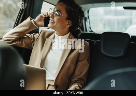 Fröhliche Frau im Business-Anzug sitzt auf dem Rücksitz ihres Autos und spricht mit dem Handy. Geschäftsfrau, die während der Fahrt mit dem Auto telefonieren. Stockfoto