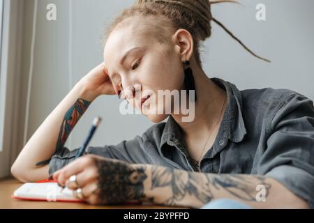 Bild von einem seriösen schönen jungen Mädchen mit Dreadlocks ein Piercing drinnen Schreiben Notizen in Notizbuch. Stockfoto