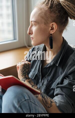 Bild von einem seriösen schönen jungen Mädchen mit Dreadlocks ein Piercing drinnen Schreiben Notizen in Notizbuch. Stockfoto